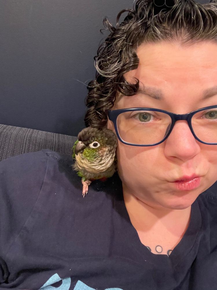 birdportant making a silly face with a small green parrot on their shoulder.