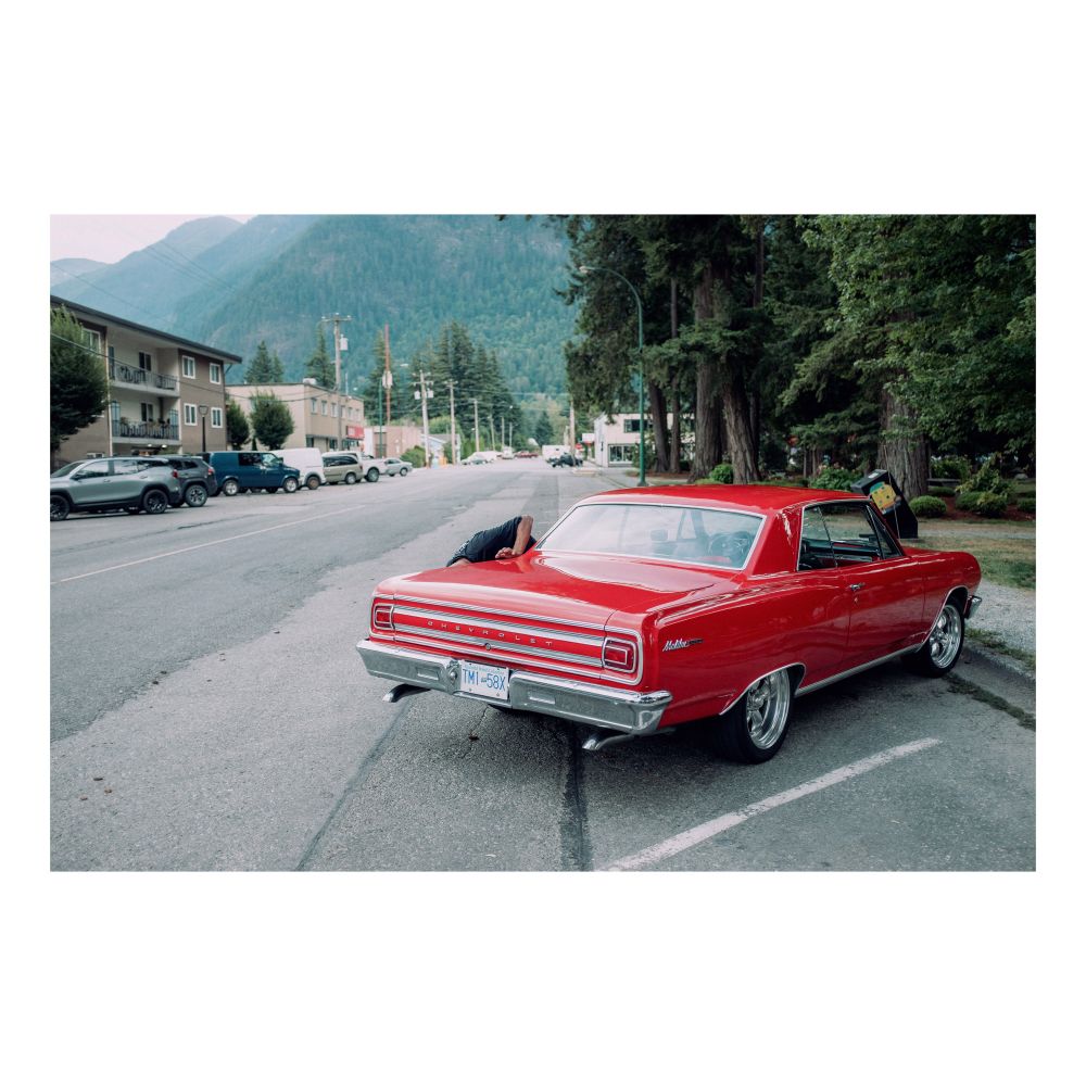 man bending down behind a vintage red chevorlet. the background contains tall trees and a mountain