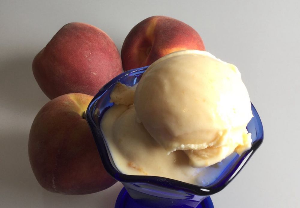 Home made buttermilk peach ice cream from a recipe in Jubilee. Two scoops of pale peach ice cream in a cobalt fluted ice cream bowl next to three unpeeled peaches onna white background. The ice cream shows a bit of melt since the recipe does not use stabilizers.