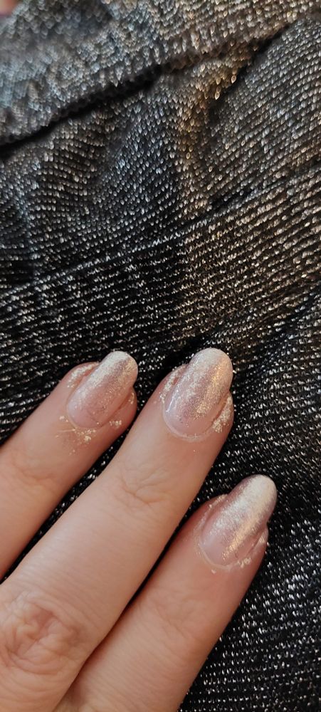 The three middle fingers of the left hand palm down on a black silvery fabric. The nails have very light pink nail polish with golden glitter polish dabbed on top.