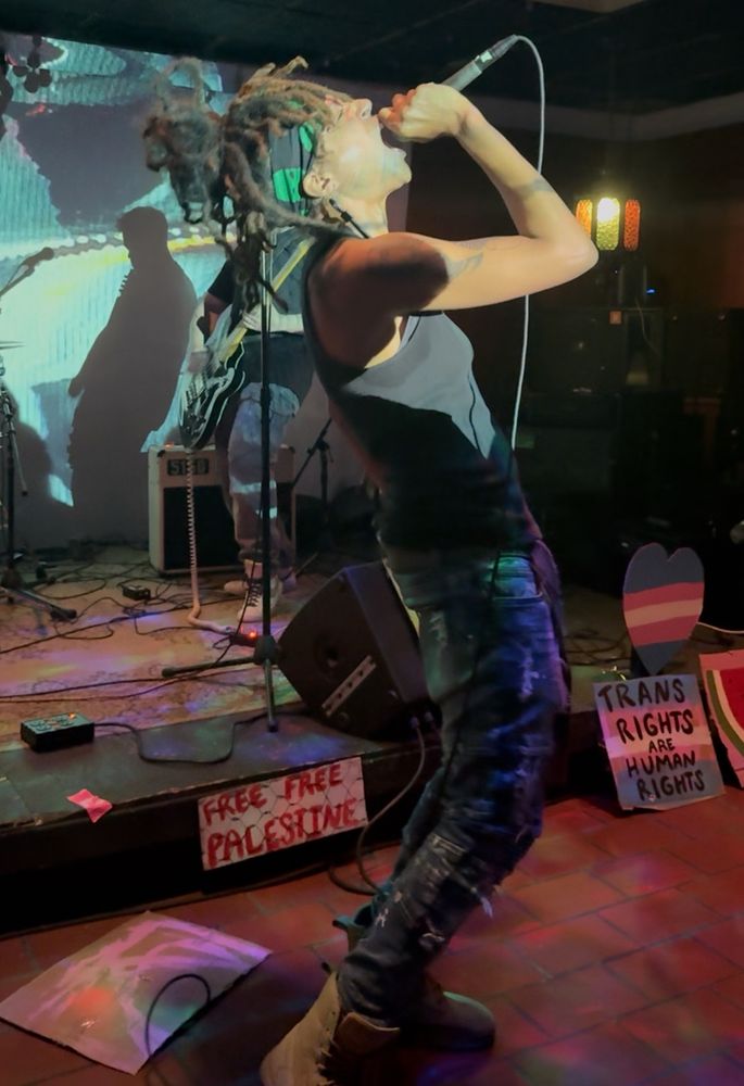 an androgynous Black woman is holding a microphone with her head tilted back, eyes shut, and mouth open wide as she screams into a microphone. her face is illuminated by a projector video and you can see various trans rights and pro palestine signs in the background 