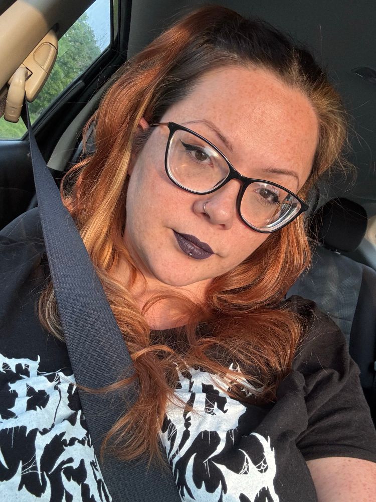 iris is a chubby redhead with lots of freckles, wearing thick-rimmed black glasses and dark purple lipstick. they have a black shirt on with white lettering, and piercings in their nose and lip.