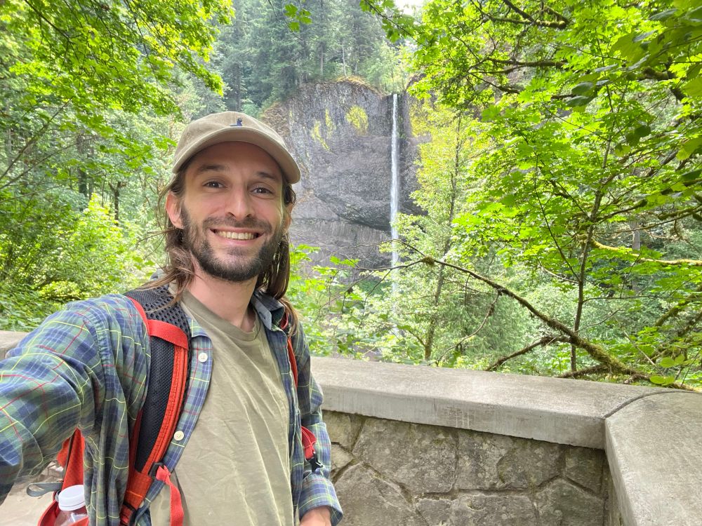 A smiling guy in a hat stands in front of a tall waterfall surrounded by greenery.