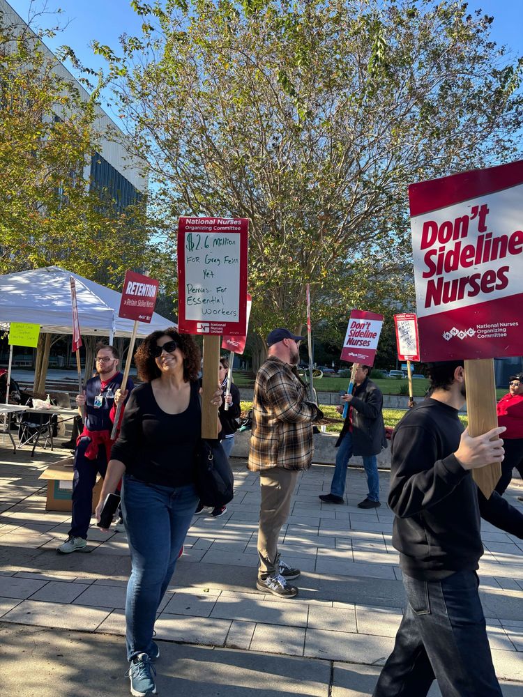 A picketer -- smiling because although striking is risky and hard and scary, it's empowering to fight for what you believe in which can be as simple and straightforward as fair pay for a day's work in your expertise and a safe workplace -- carrying a sign that says "$2.6 million for Greg Feirn yet pennies for essential workers!" ✊