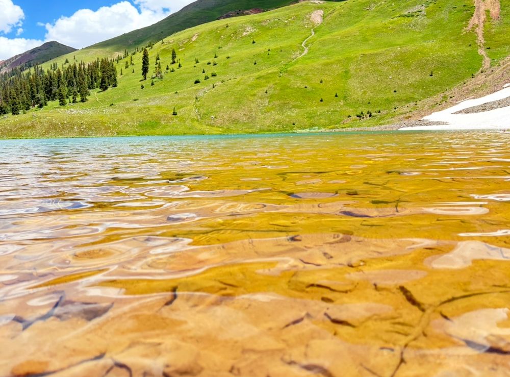 Velocity Basin, near Silverton Colorado . Across the water shot from water surface. The water is very clear and the tan colored rocks are visible through the water. The mountain in the background is green and healthy looking. A patch of leftover snow in the background.