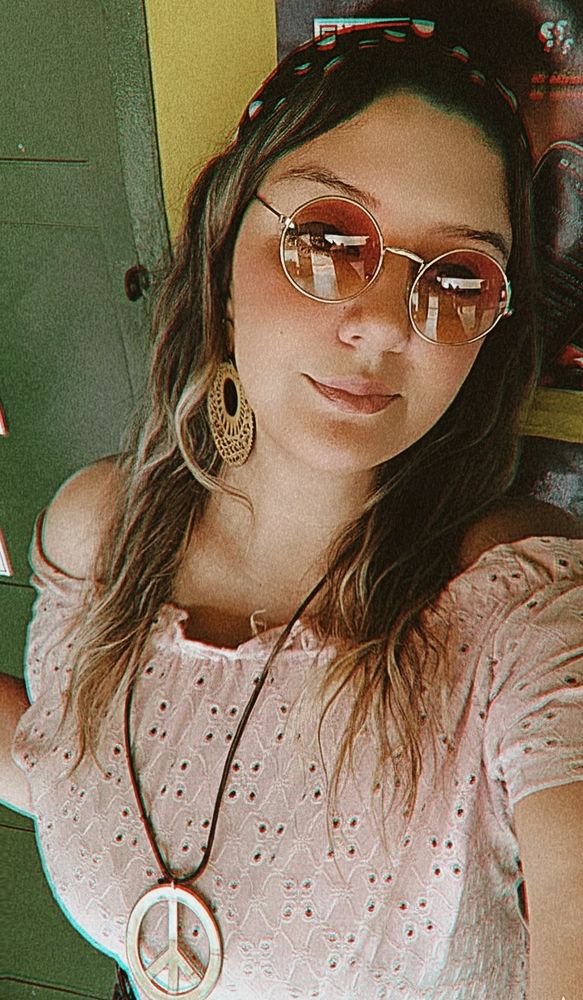 Ela usa uma blusa ombro a ombro cor-de-rosa clara com detalhes bordados, um colar grande com o símbolo da paz, e brincos grandes de madeira trabalhados em formato rendado. Também usa óculos redondos de lente rosada e uma faixa estampada no cabelo, que está solto, com ondas suaves e pontas mais claras. A expressão do rosto é serena e é possível ver uma porta verde e parte de um poster visível. A foto tem um filtro retrô, com tons quentes e um leve efeito de distorção de cores, o que reforça o clima vintage da imagem.