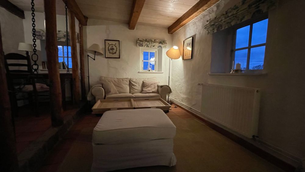 Eine gemütliche Sofaecke in einer Zimmerecke mit kleinen Fenstern, durch die blaues Dämmerlicht fällt, viele Holzbalken, Tischchen, Bilder, Lampen. Sehr altes Haus.