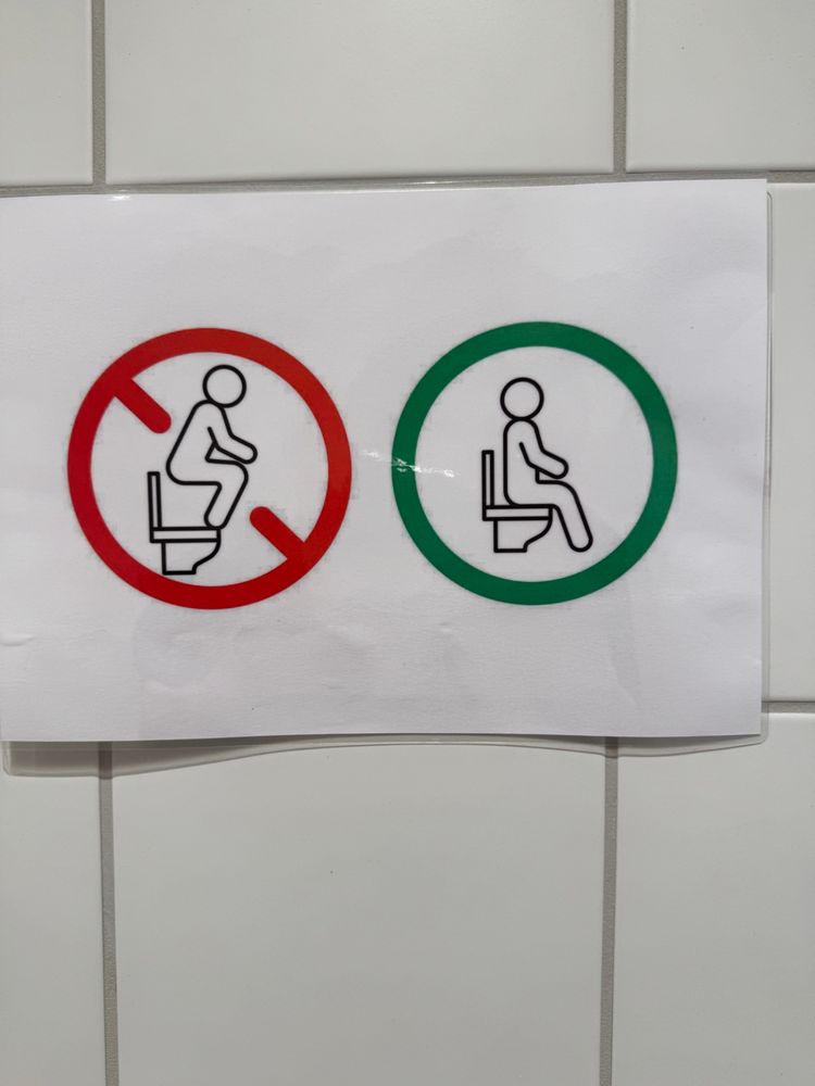 Auf dem Bild ist ein Schild an einer gekachelten Wand. Links hockt eine Figur auf einer stilisierten Toilette und ist rot eingekreist und rot durchgestrichen. Daneben Sitz eine Figur auf der Toilette und ist grün eingekreist.