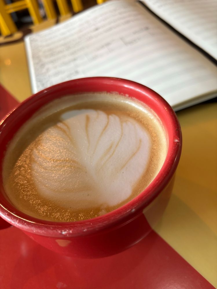 A latte in a red cup. The foam art on top is in the shape of a leaf. Behind the latte is an open music manuscript notebook. 