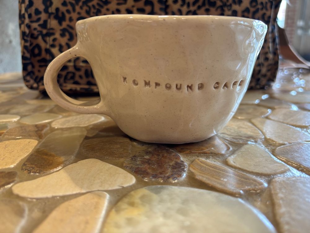 A coffee mug sits on a table in a coffee shop. The name of the shop, Kompound Cafe, is stamped into the surface of the mug. 