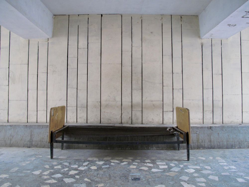 Ein Bettgestell steht mittig unter einer Brücke, im Vordergrund Betonboden und an der Wand dahinter senkrechte Streifen.