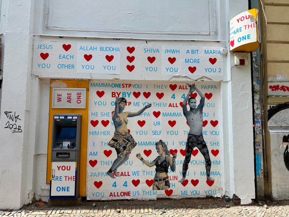 Links ein Geldautomat rechts daneben und darüber viele Plakate mit roten Herzen und Sprüchen wie Love each other, darauf drei tanzende Personen.
