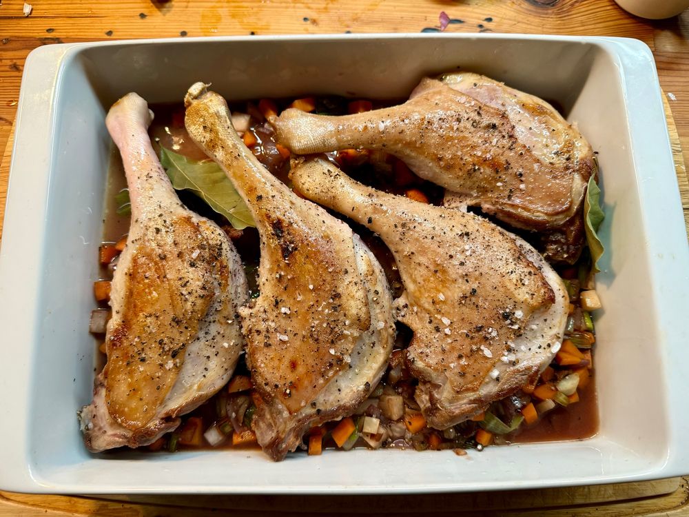 ChatGPT: Ein köstlich angerichtetes Quartett aus angebratenen Entenkeulen liegt in einer Auflaufform auf einem Bett aus gewürfeltem Gemüse und Brühe. Grobes Salz, Pfeffer und Lorbeerblätter verleihen dem Gericht eine rustikale, aromatische Note.