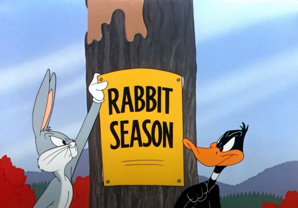 Sign hanging from a tree says rabbit season. But Bugs Bunny is about to tear it down.