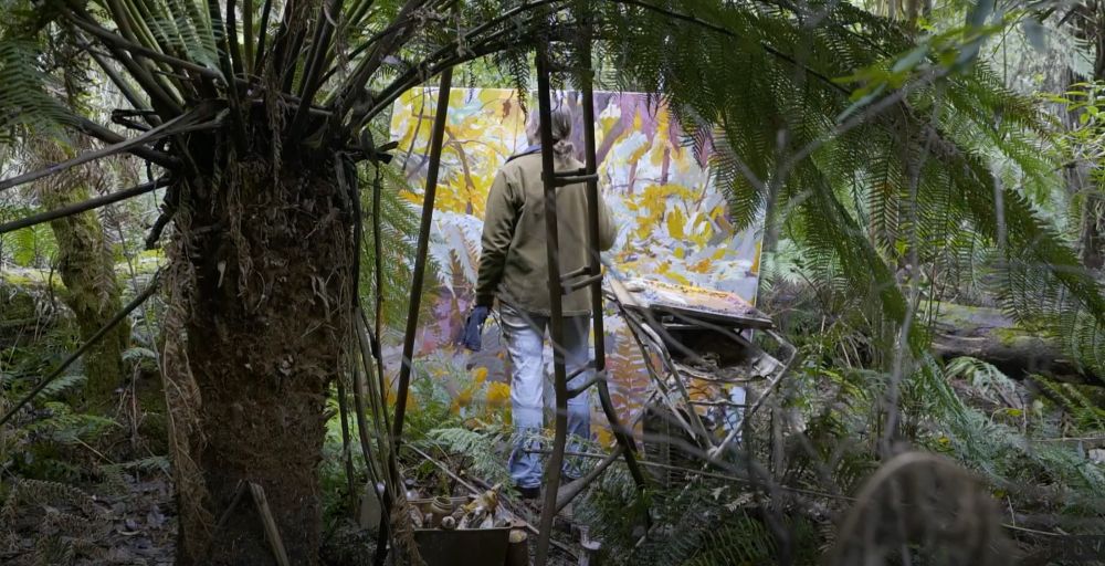 A photo of Mary Tonkin, painting on a canvas that is as big as her out in the middle of the woods