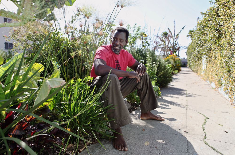 Ron Finley, a barefoot Black man wearing casual clothes, sitting on a sidewalk surrounded by lush plants. 