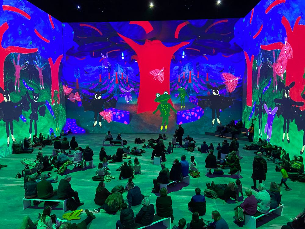 Crowd of People sitting on the floor surrounded by an Immersive installation of a David Hockney artwork projected on tall screens all around them. The artwork is of a surreal forest scape using Fauvist colour- red tree trunks with blue leaves and a green floor