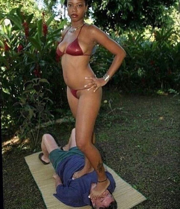 uma mulher de biquíni de pé pisando em um homem deitado em uma esteira num jardim 