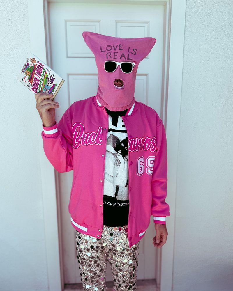 buckaroos chuck tingle holding a copy of LUCKY DAY in front of a white wall and door. he is wearing pink bag over head with matching varsity jacket that says BUCKAROOS number 69 and sequin silver pants