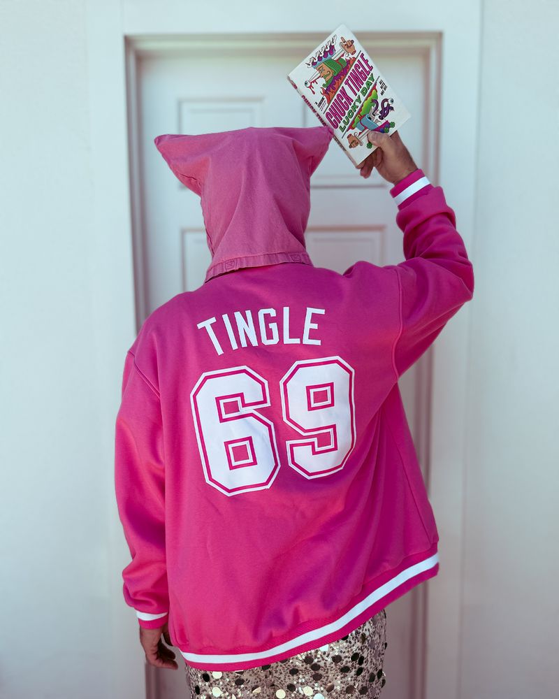buckaroos chuck tingle holding a copy of LUCKY DAY in front of a white wall and door. he is facing away, wearing pink bag over head with matching varsity jacket. it is the back of the jacket and it says TINGLE number 69