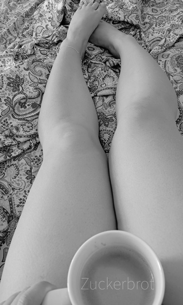 Beine einer Person, die auf einem gemusterten Bett liegen, mit einer Tasse Kaffee auf den Oberschenkeln und am linken Knöchel ein Fußkettchen