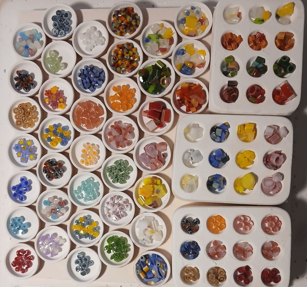 kiln shelf half covered in little dishes full of murrine, half of which has been carefully arranged and half of which is just willy nilly. also 3x 12 piece round 1 inch molds, similar ratio.