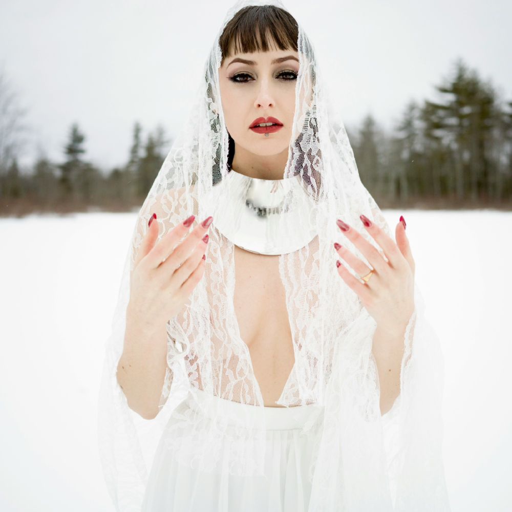 A woman with red lipstick, roles with a long white skirt stands hands raised on a snowy white scene on a frozen lake