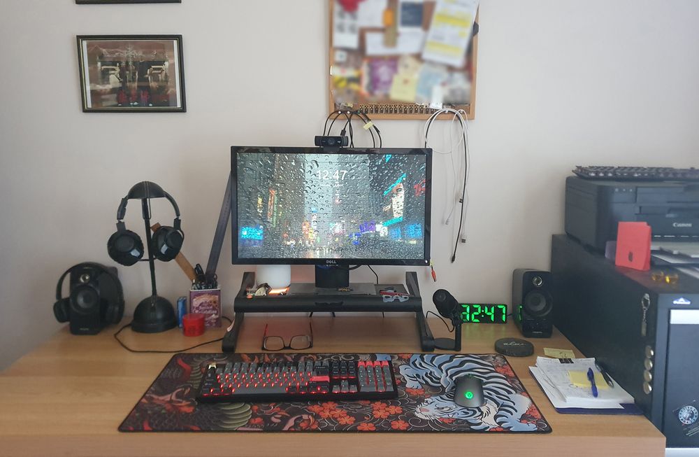 shot of my desk showing a monitor on a stand, blurred out pinboard behind it (because private, you sneaks!). There's a lamp, bluetooth headset, keyboard and mouse on an XK gaming mat, speakers, clock, mic, note paper and pen. You can see the edge of a filing cabinet with a printer on it to the right.