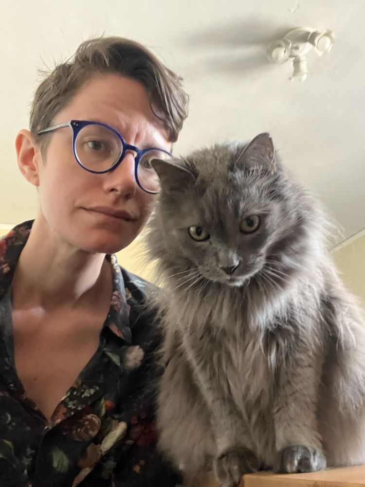 A selfie of a person and a cat looking very seriously into the camera. The person has short brown hair, round blue glasses, and a dark floral shirt. The cat is supremely grey and fluffy.