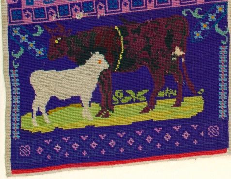 A detail of the cows from the sampler, now right side up. A dark brown-purple cow stands with a white calf on bright green and yellow grass against a dark blue-purple background, surrounded by light blue and pink geometric and floral borders. 