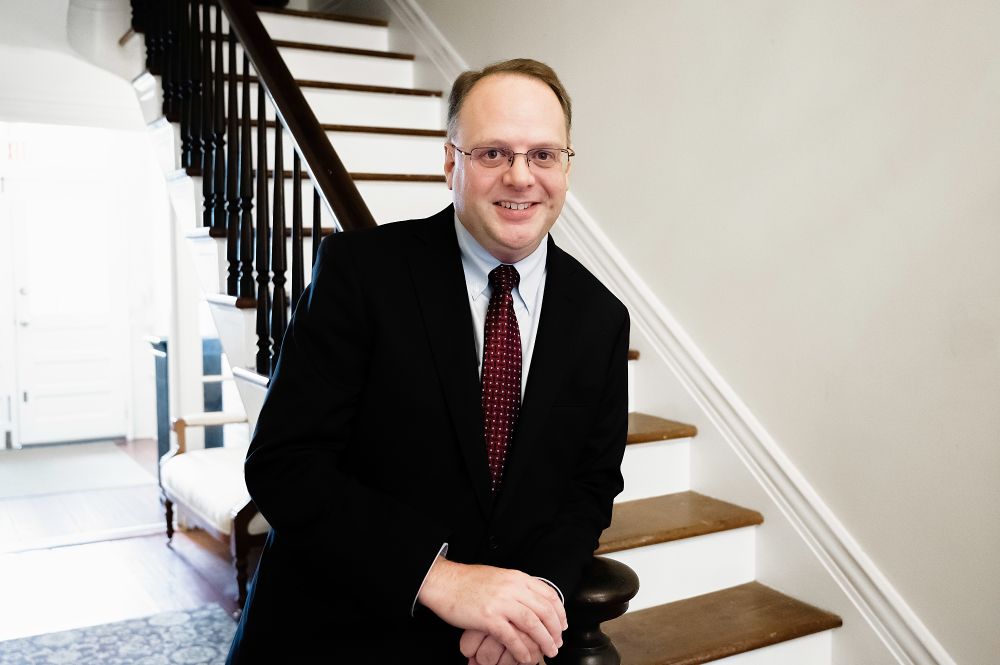Photo of Michael Thrasher in black suit with red tie in front of a staircase. 