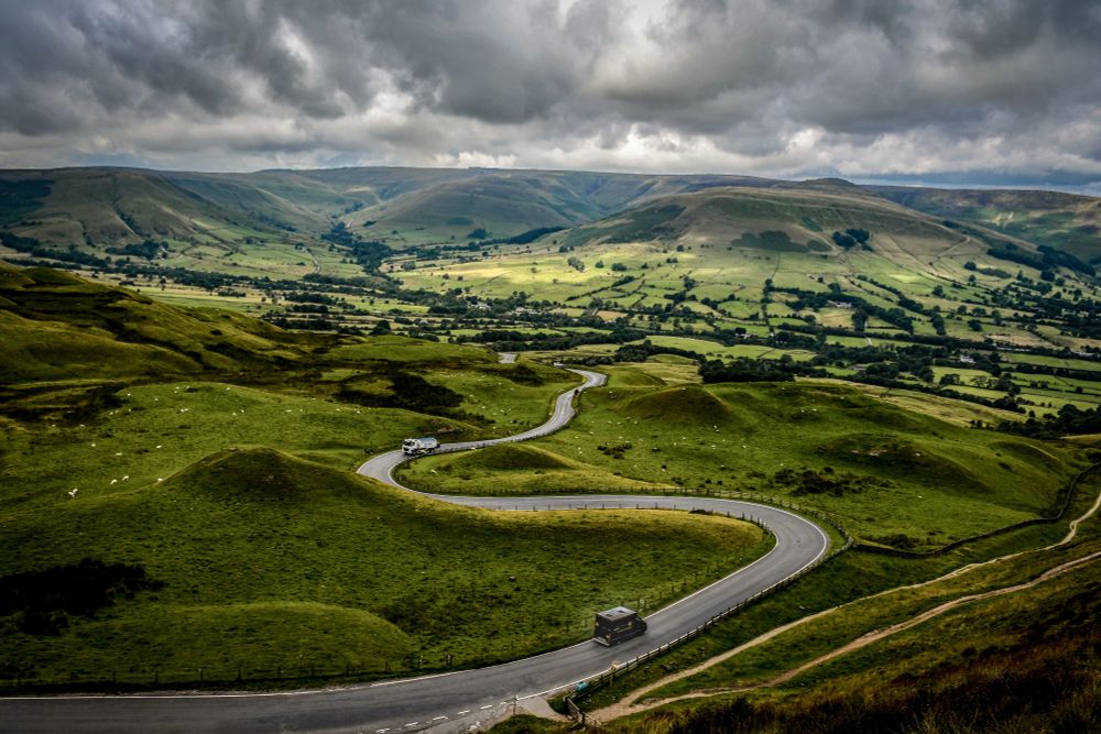 A van makes it's way along the winding road from the bottom of Mam Tor, Derbyshire. Light pierces cloud to illuminate the fells to which the road leads.