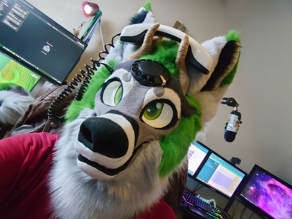 A selfie of me in a baggy red shirt wearing a grey canine fursuit head with green highlights and a rotary encoder from a phone embedded into the forehead and a phone handset being held by pair of antlers and a cable going from the handset into the side of the head, taken in front of some screens.