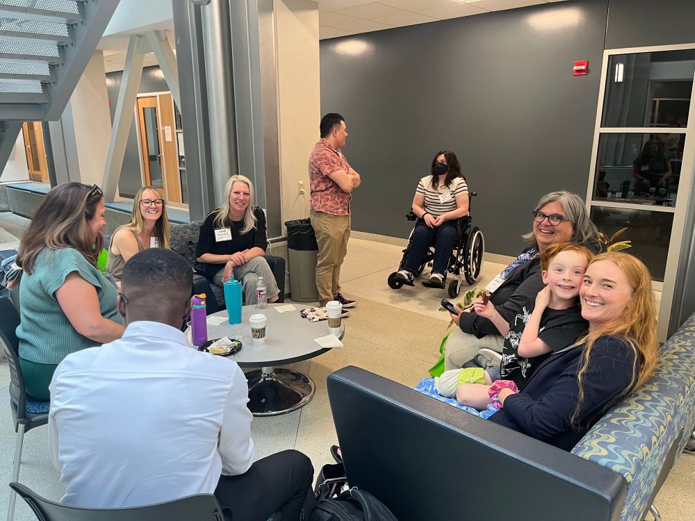 Group photo of in-person attendees gathered in a circle. Attendees are sitting on two couches with a round table in the middle. Group includes Michelle Eble, Erin Clark, her son, Sarah Warren-Riley, Angela Haas, victoria braegger, and Derek Sparby.