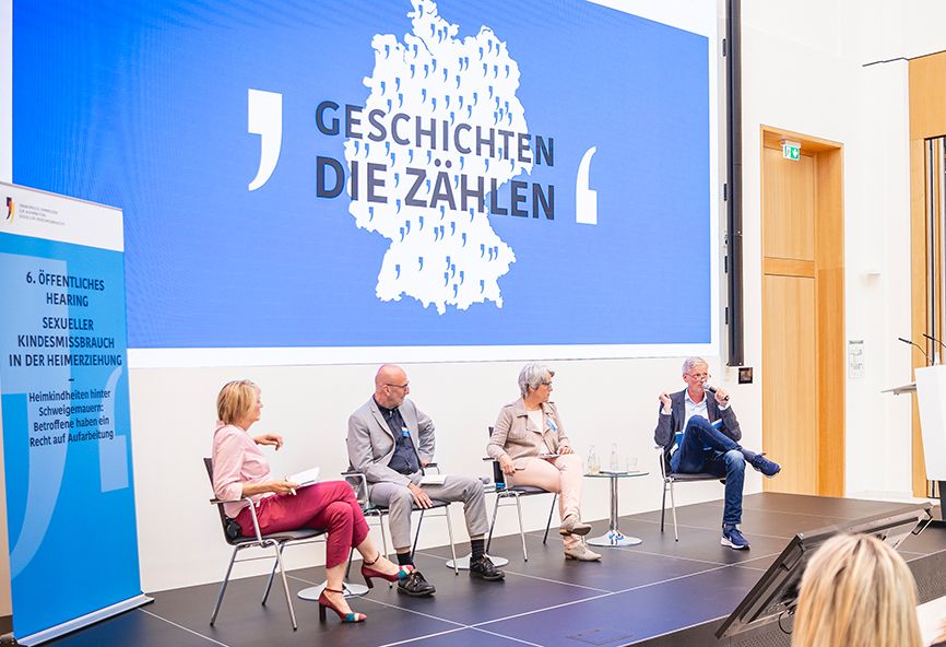 Bühne mit vier Personen, vom linken Bühnenrand fotografiert. Zu sehen sind auf Stühlen von links nach rechts: Anke Plättner, Ignaz Raab, Kerstin Claus, Wolfgang Schröer. Im Hintergrund Logo der Aufarbeitungskommission.