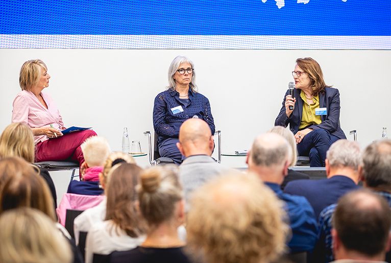 Aus der Ferne ist eine Bühne mit drei Frauen zu sehen, eine von ihnen hält ein Mikrofon. Im Vordergrund die Köpfe der Zuhörenden.