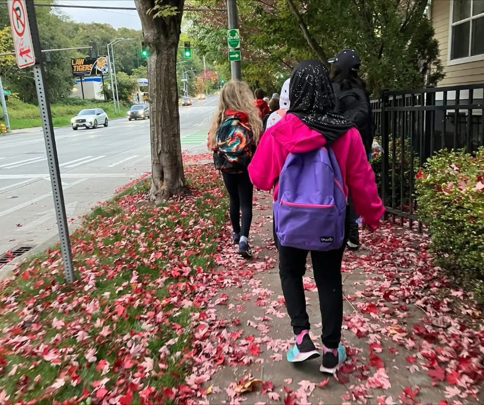 Three children with backpacks, walking along a sidewalk covered with red autumn leaves, near a street intersection with traffic signs.
