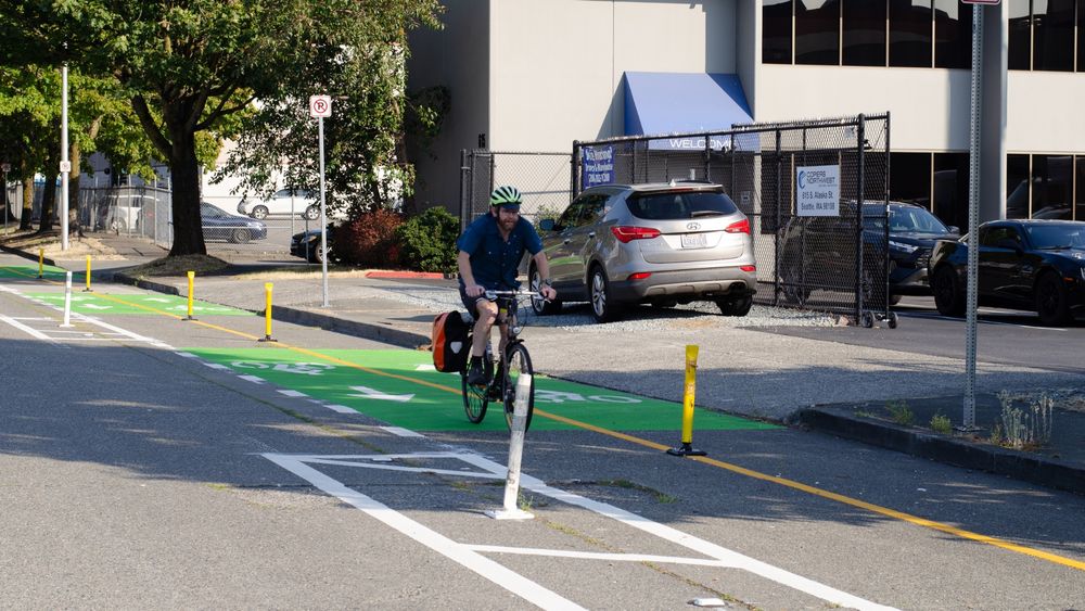 A cyclist wearing a helmet and carrying a side bag rides along a designated green bike lane on a sunny day, with parked cars and a building in the background. 