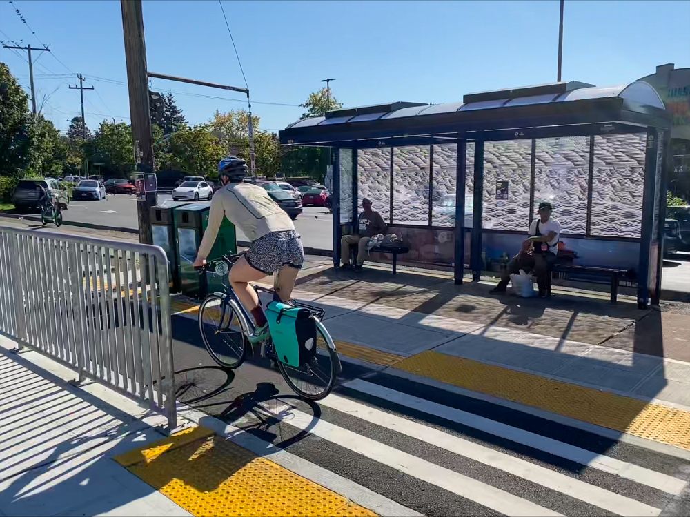 A person bikes as people sit and wait for the bus.