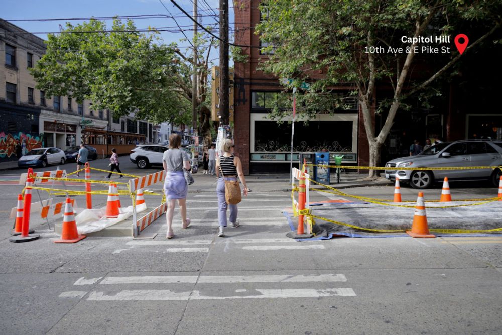 Two people cross the street at 10th Ave and E Pike St in a crosswalk. There is construction signage, caution tape, and traffic cones surrounding two new concrete median islands along the center of the street.