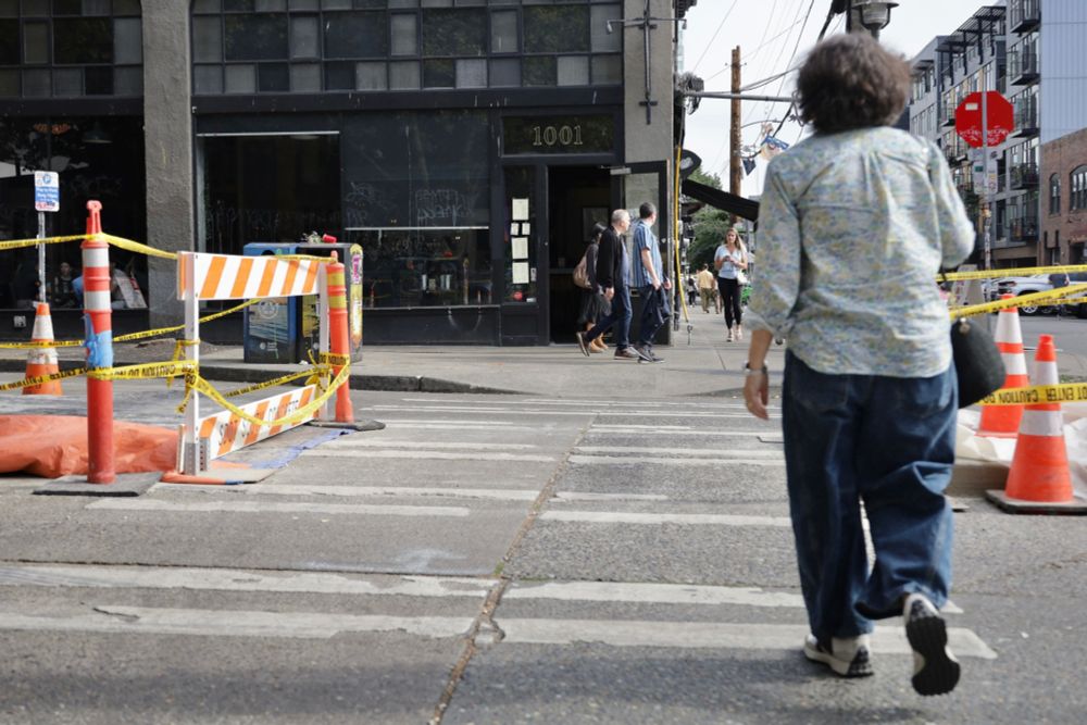 A person crosses the street at 10th Ave and E Pike St in a crosswalk. There is construction signage, caution tape, and traffic cones surrounding two new concrete median islands along the center of the street.