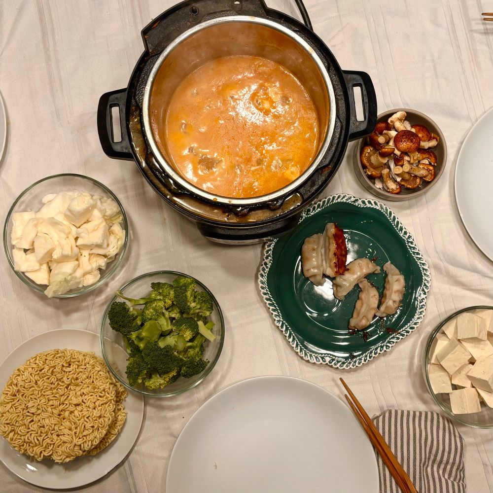 A gorgeous photo of an InstantPot filled with a simmering vegan tonkatsu broth, surrounded by bowls of broccoli, tofu, mushrooms, cabbage, ramen noodles and vegetable dumplings.
