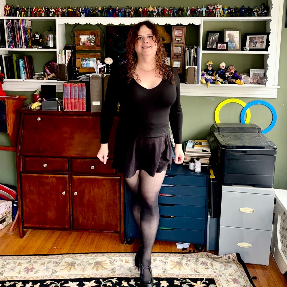 Jadzia is wearing a silver necklace with a small silver star charm, a black long-sleeve low-cut shirt, a short black skirt, sheer black tights, and black Fluevog heels.