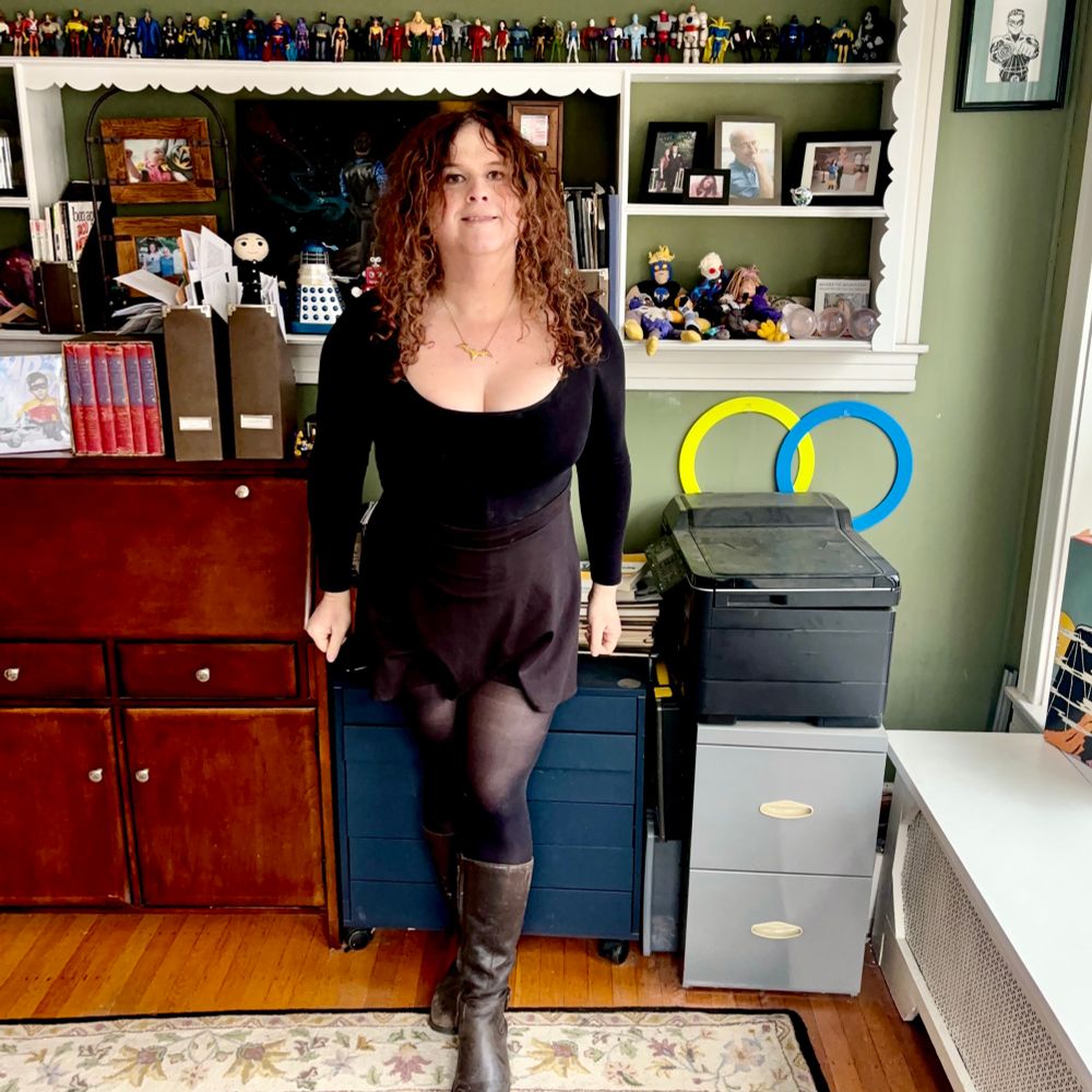 Jadzia is wearing a gold necklace shaped like a batarang, a low-cut long-sleeve black bodysuit, a short black skirt, black tights, and high black boots.