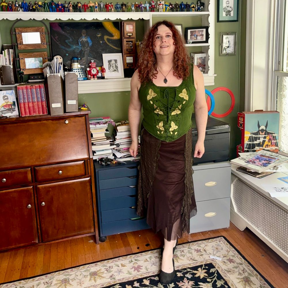 Jadzia is wearing an necklace with a blue stone in an ornate setting, a green bodice with gold embroidered leaves, a long brown skirt made of different layers and textures of fabric, and black Sketchers ballet flats.