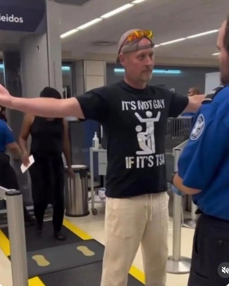 Man being patted down by TSA wearing a shirt that says “It’s not gay if it’s TSA” 