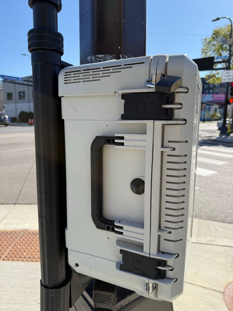 A grey plastic box with vent holes and the word miovision attached to a traffic pole. 