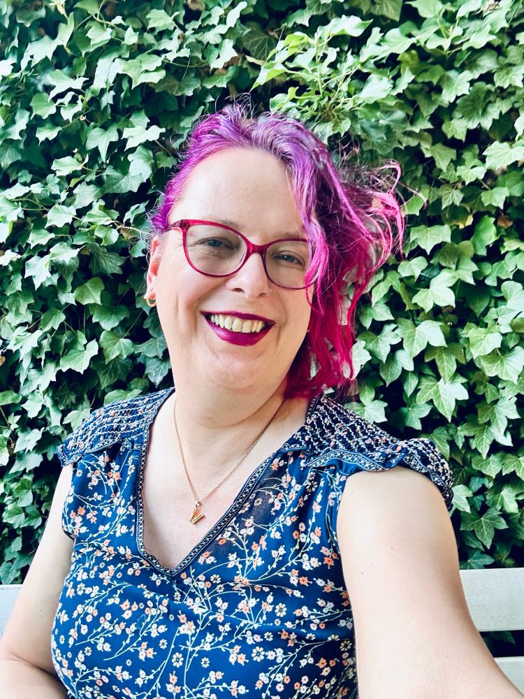 Selfie of me smiling into the camera with asymmetric, purple hair, matching purple lipstick and glasses, in a navy summer dress with small flower ornaments, a golden necklace with a V pendant sitting on a bench in front of an ivy-covered wall