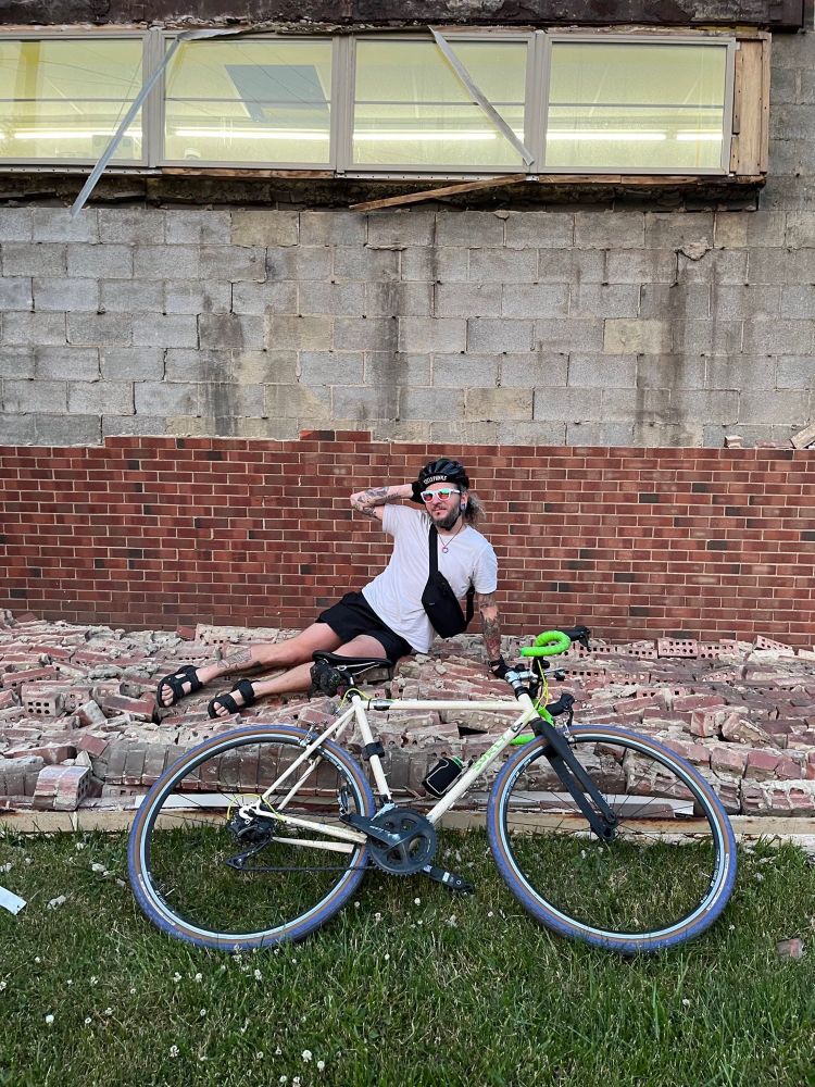 Ee lounges in a bike helmet, white t-shirt, short shorts, sandals, and a fashionable set of rose colored glasses on a carpet of bricks. In the foreground is a layed down bicycle. In the background is a cinder block wall, the top 2/3rds are barren of brick while a precious couple dozen layers still remain affixed to the block.