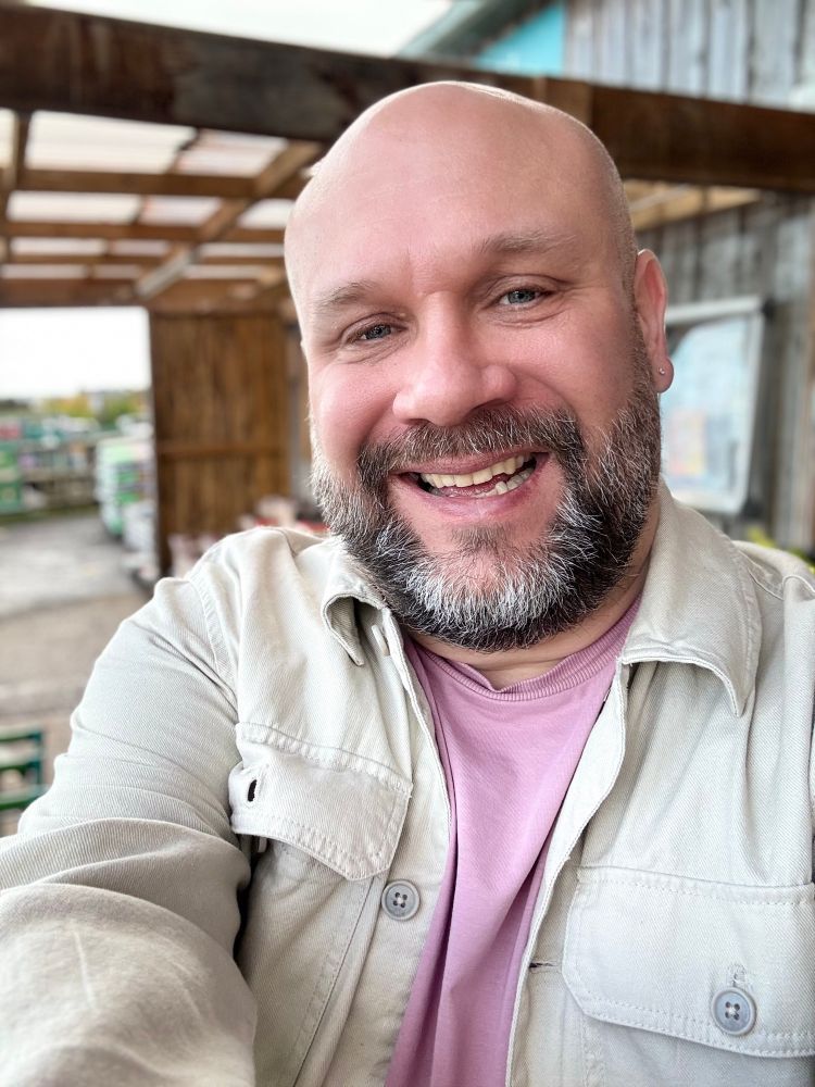 A smiling man with a beard and shaved head takes a selfie under a wooden canopy. He’s wearing a light beige overshirt and a mauve T-shirt.